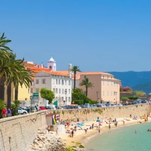 Plage d’Ajaccio animée par des vacanciers sous un ciel bleu, bordée de bâtiments aux façades pastel et de palmiers majestueux. 